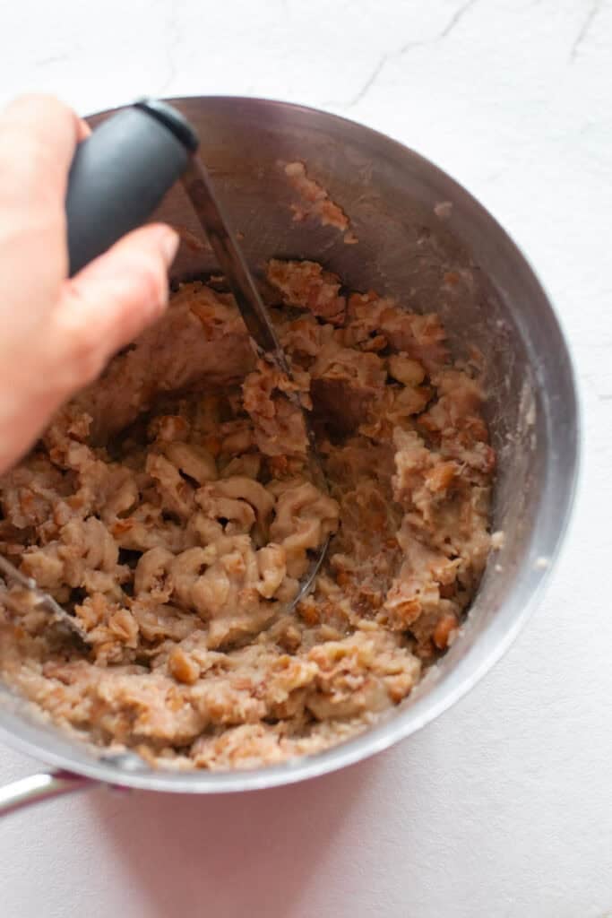 Mashed pinto beans in a sauce pan with a potato masher.