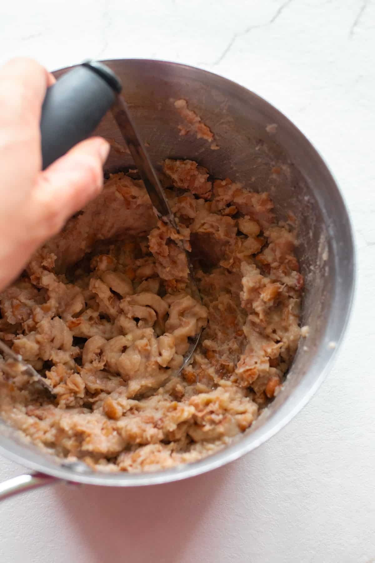 Mashed pinto beans in a sauce pan with a potato masher.