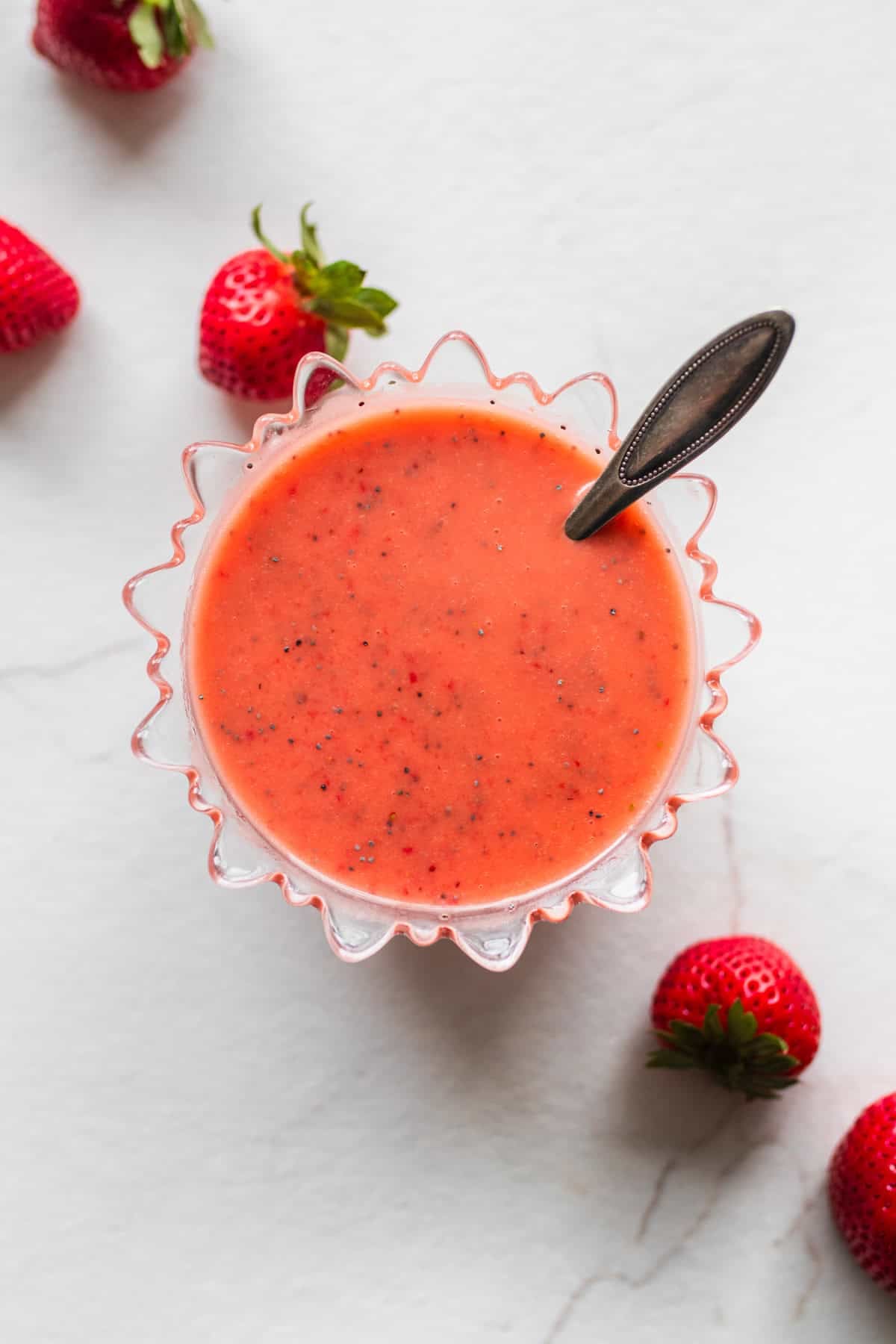 A bowl of strawberry salad dressing with a spoon in it and strawberries on the side on a counter top.