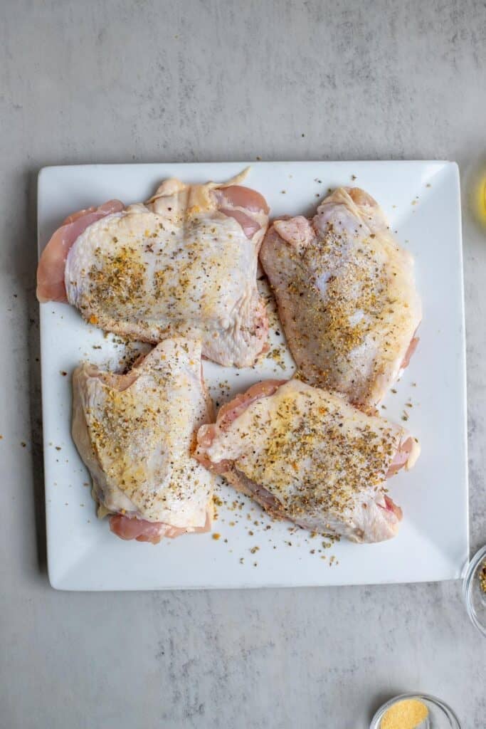 Four chicken thighs on a white platter seasoned with salt, pepper, and Italian seasoning.