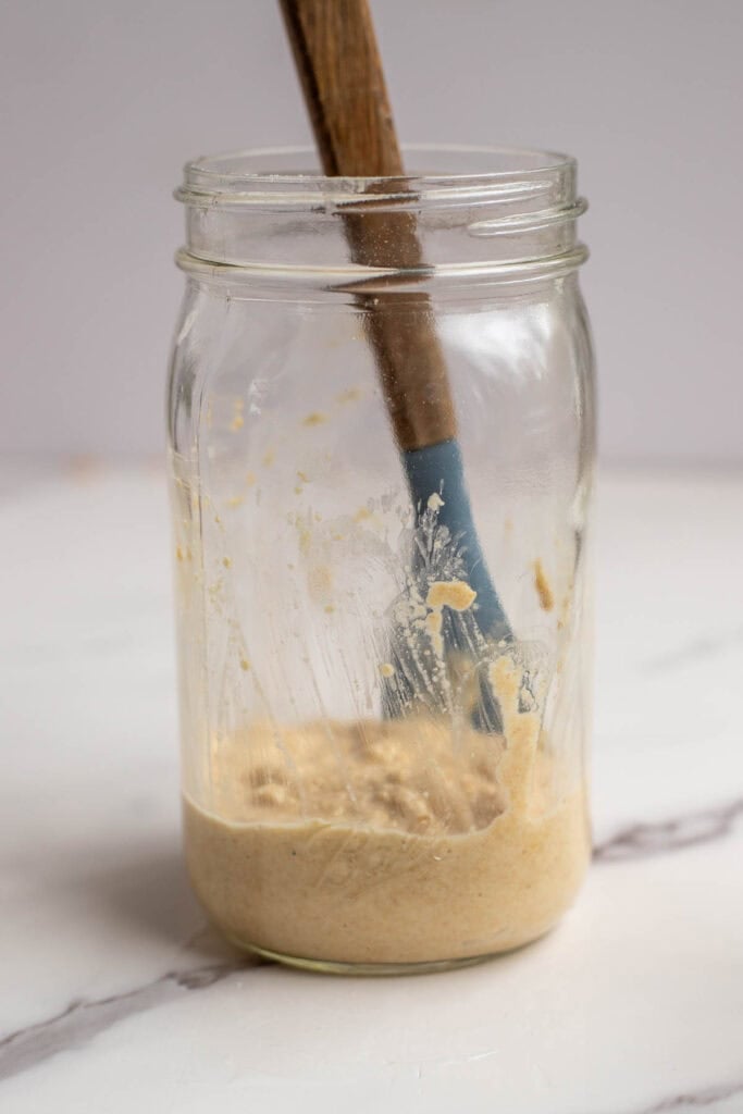 Using a silicone spatula to scrape down the sourdough starter from the sides of a mason jar.