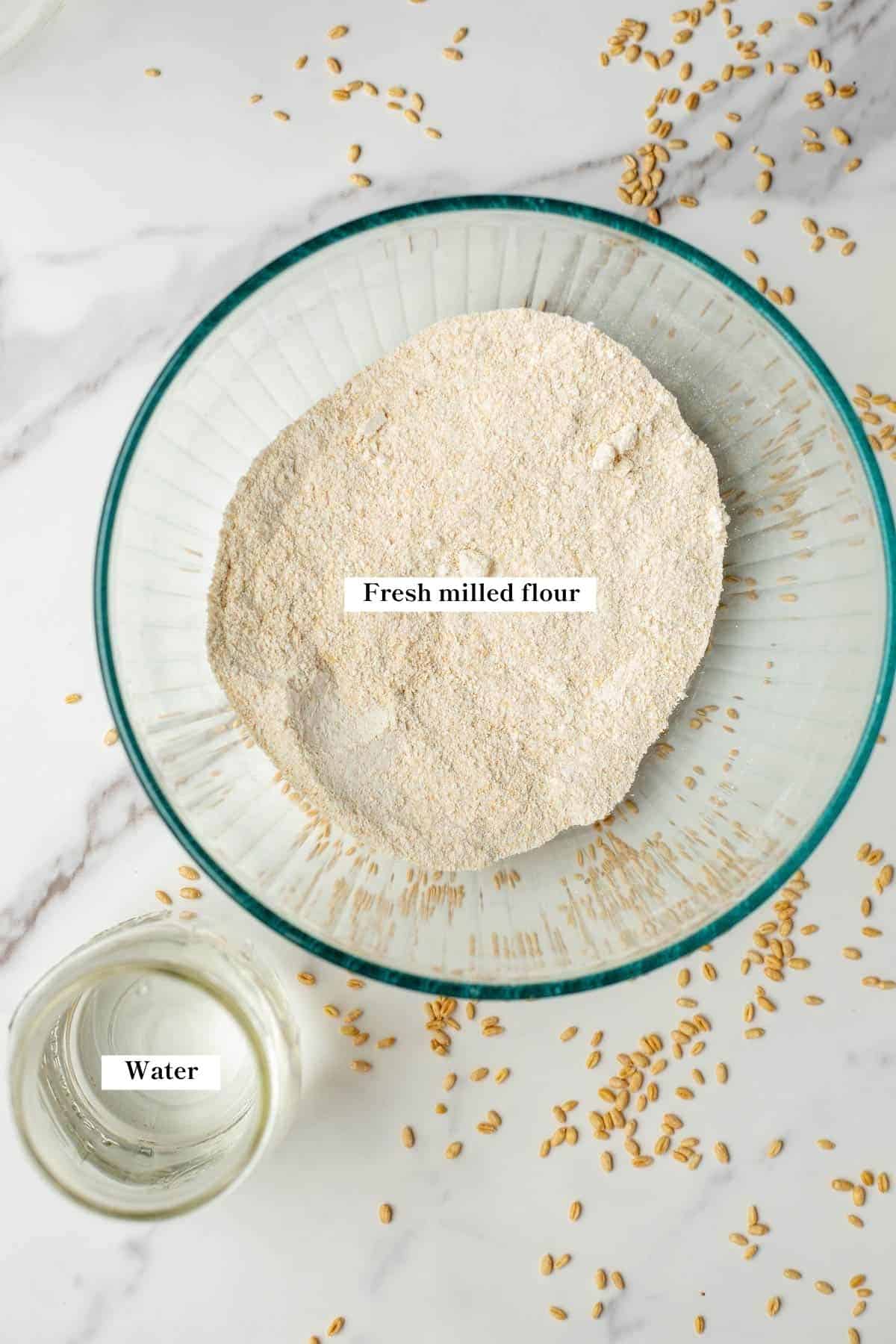 a glass bowl full of freshly milled flour and a cup of water beside it.