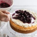 A hand spooning cherries on top of a cheesecake.
