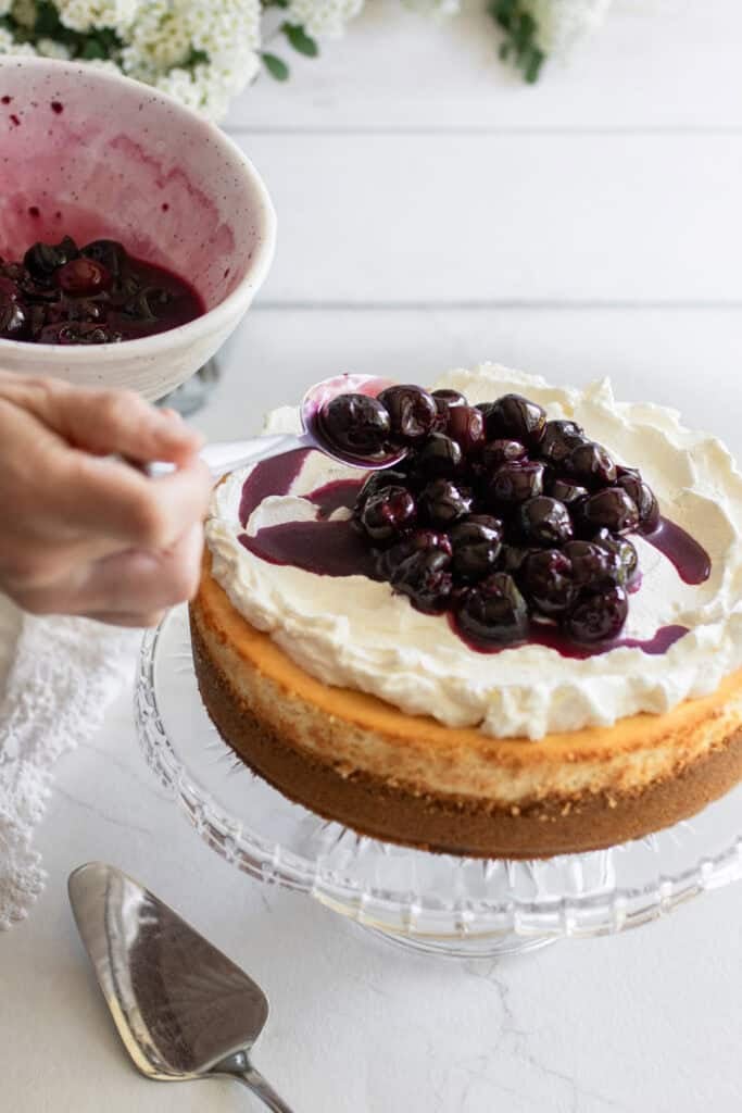 A hand spooning cherries on top of a cheesecake.
