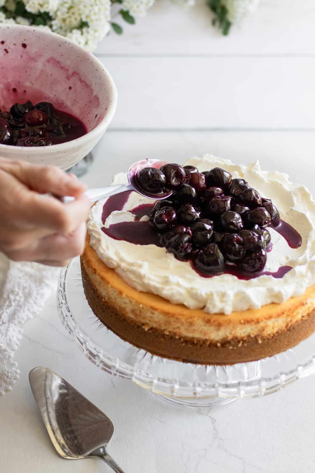 A hand spooning cherries on top of a cheesecake.