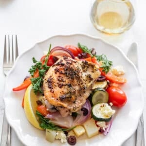 A plate with a chicken thigh and veggies and a knife and a fork on either side.