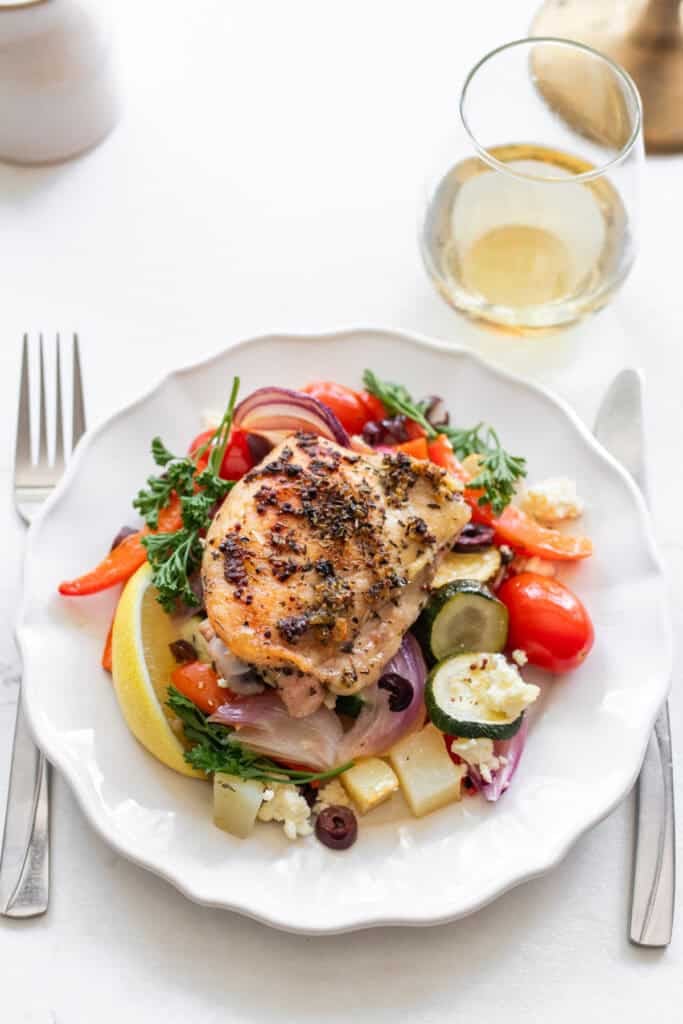 A plate with a chicken thigh and veggies and a knife and a fork on either side.