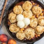a cast iron skillet full of peach cobbler topped with three scoops of vanilla ice cream.