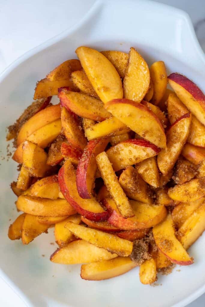 Sliced peaches tossed with brown sugar in a white mixing bowl.