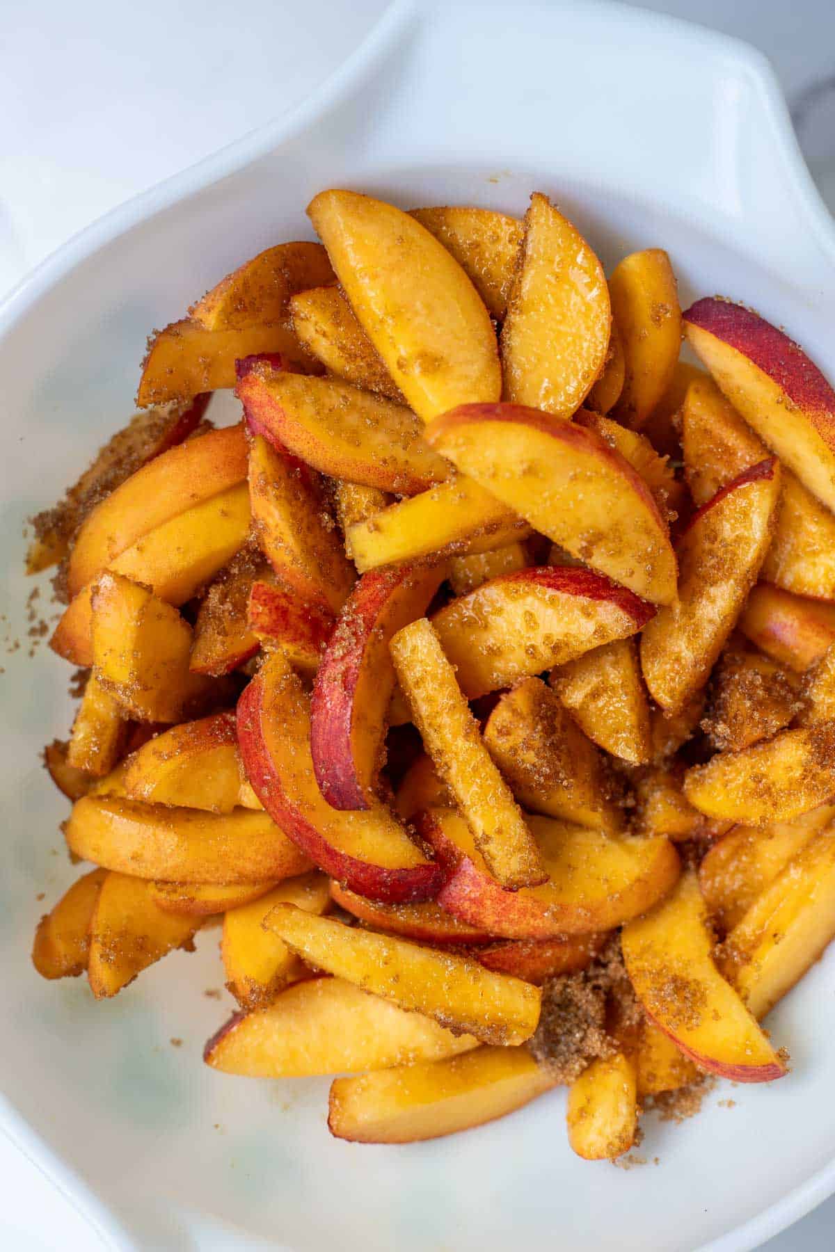 Sliced peaches tossed with brown sugar in a white mixing bowl.
