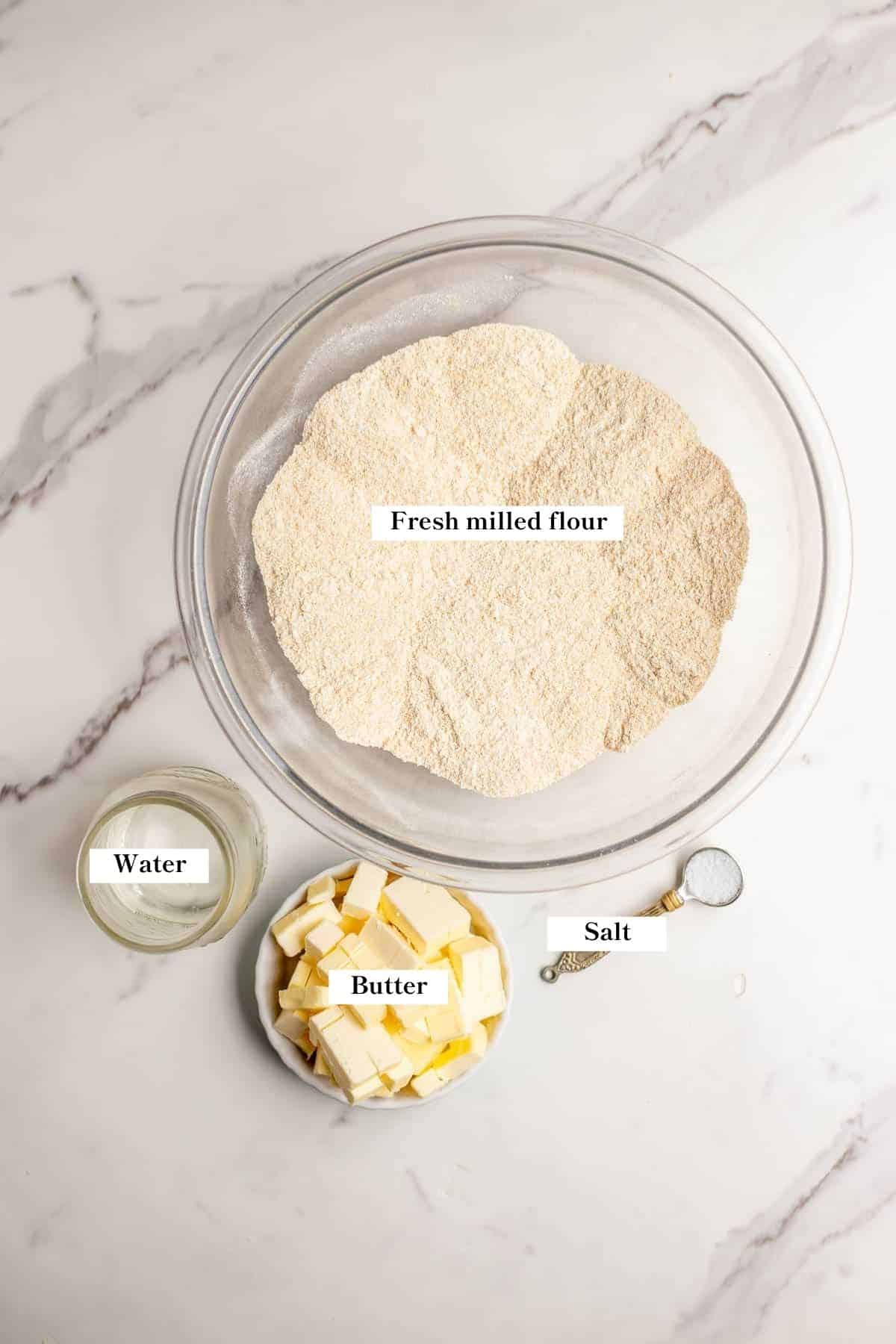 Freshly milled flour, cubed butter, water, and salt on a marble countertop.