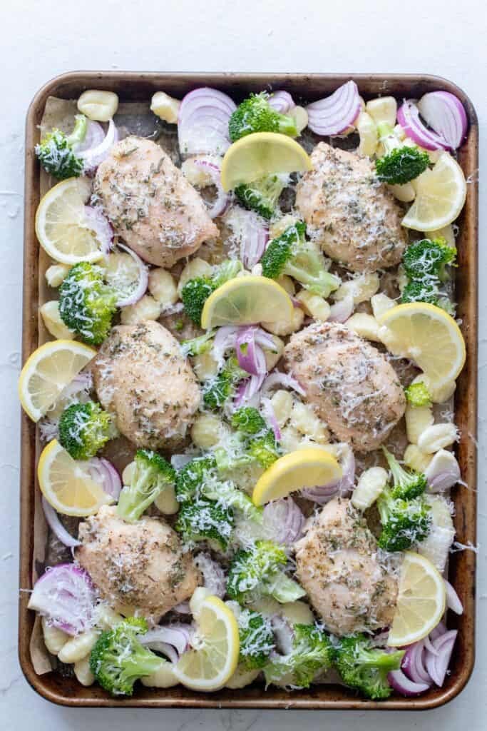 Sheet pan of chicken breasts with veggies, gnocchi and lemon slices before roasting.