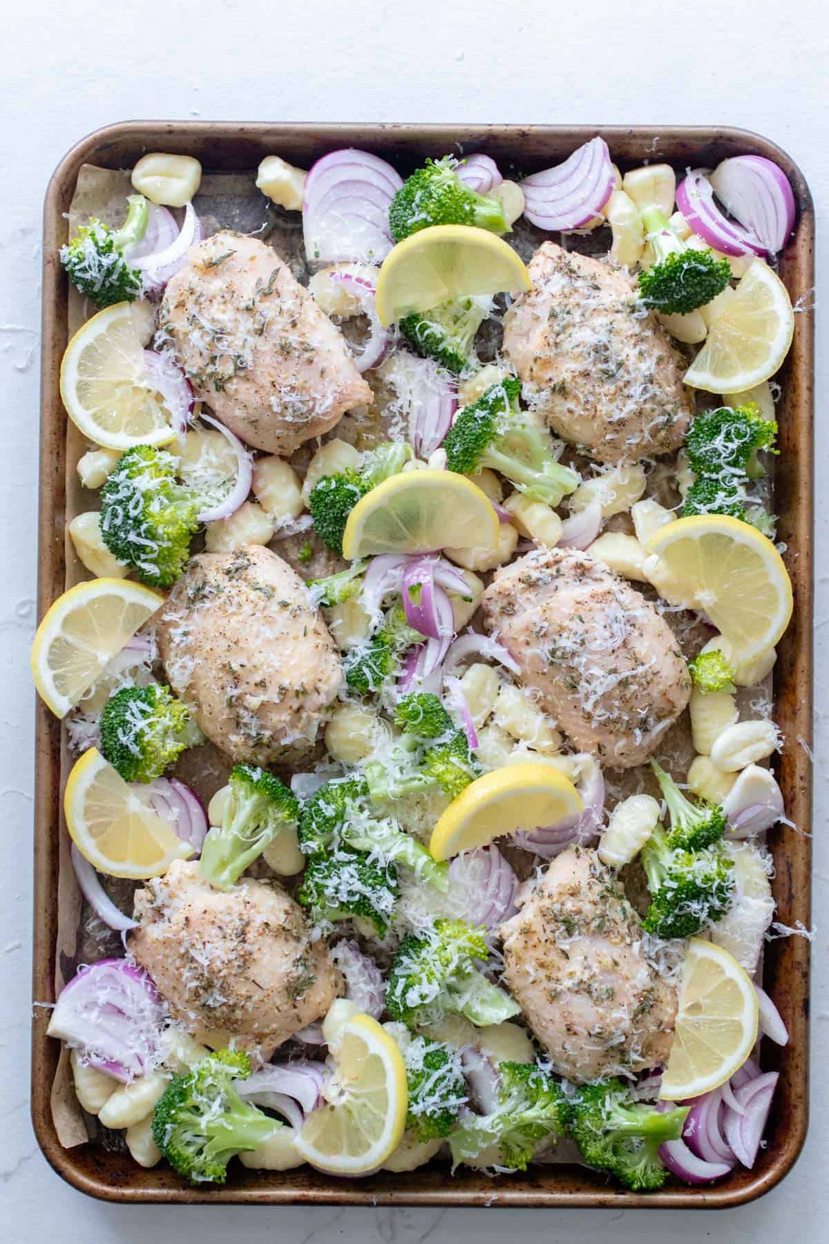 Sheet pan of chicken breasts with veggies, gnocchi and lemon slices before roasting.