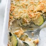 A large silver spoon scooping out zucchini gratin from a casserole dish.