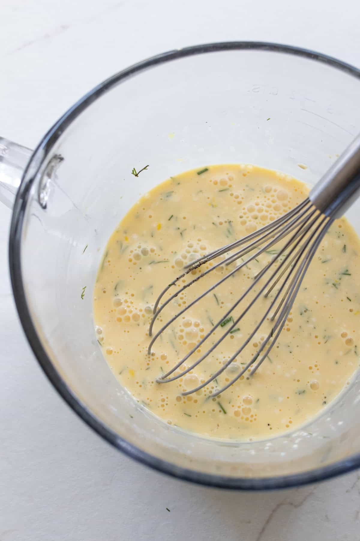 Eggs and other ingredients in a large glass measuring bowl with a whisk.