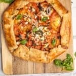 tomato galette on a cutting board with thyme, basil, and tomatoes surrounding it.