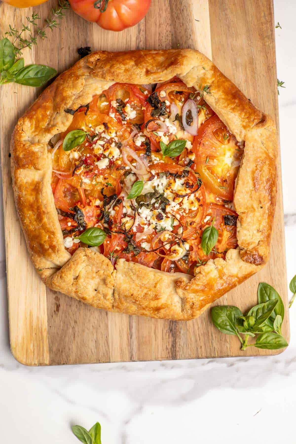 tomato galette on a cutting board with thyme, basil, and tomatoes surrounding it.