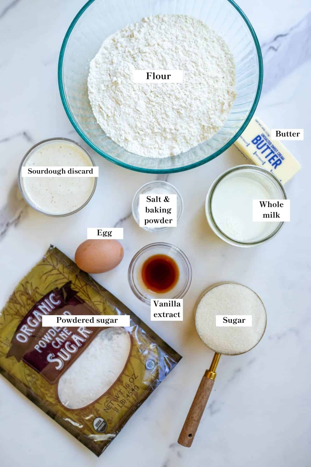 Ingredients for baked sourdough donuts in bowls, measuring spoons, and bags laid out on a white marble countertop. 