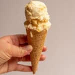 A hand holding a small ice cream cone with a scoop of peach ice cream on top.
