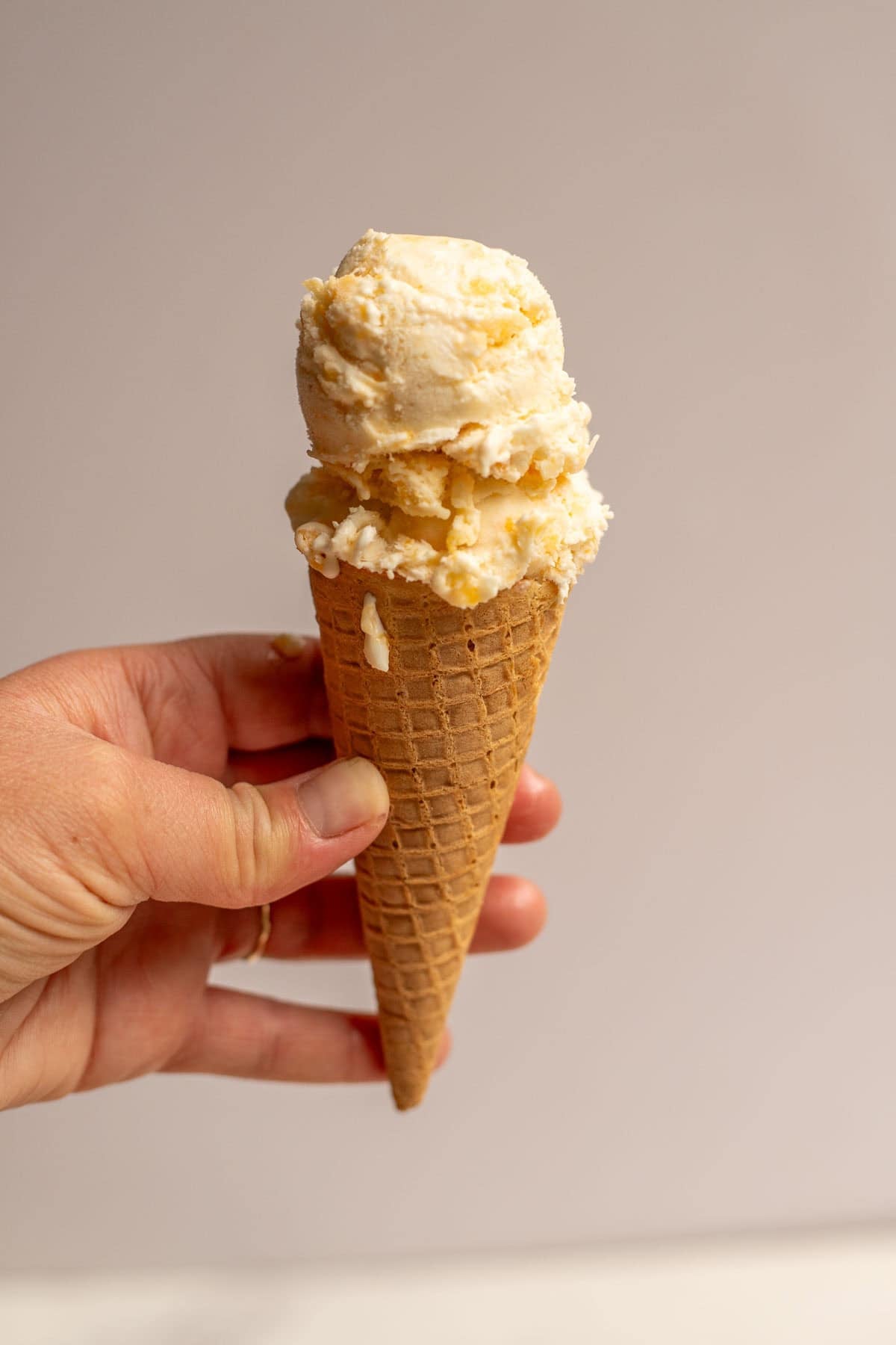A hand holding a small ice cream cone with a scoop of peach ice cream on top.
