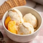 a white bowl with three scoops of peach ice cream and fresh peaches on the side.