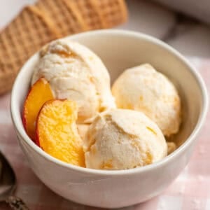 a white bowl with three scoops of peach ice cream and fresh peaches on the side.