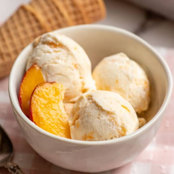 a white bowl with three scoops of peach ice cream and fresh peaches on the side.