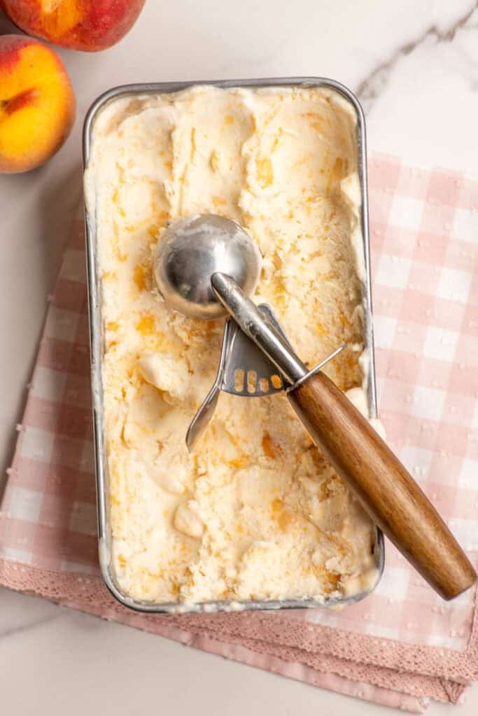 An ice cream scoop placed on top of a loaf tin full of peach ice cream.