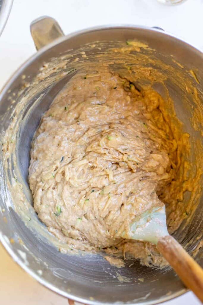 A spatula folding in dry ingredients into a zucchini bread batter in a stand mixer bowl.
