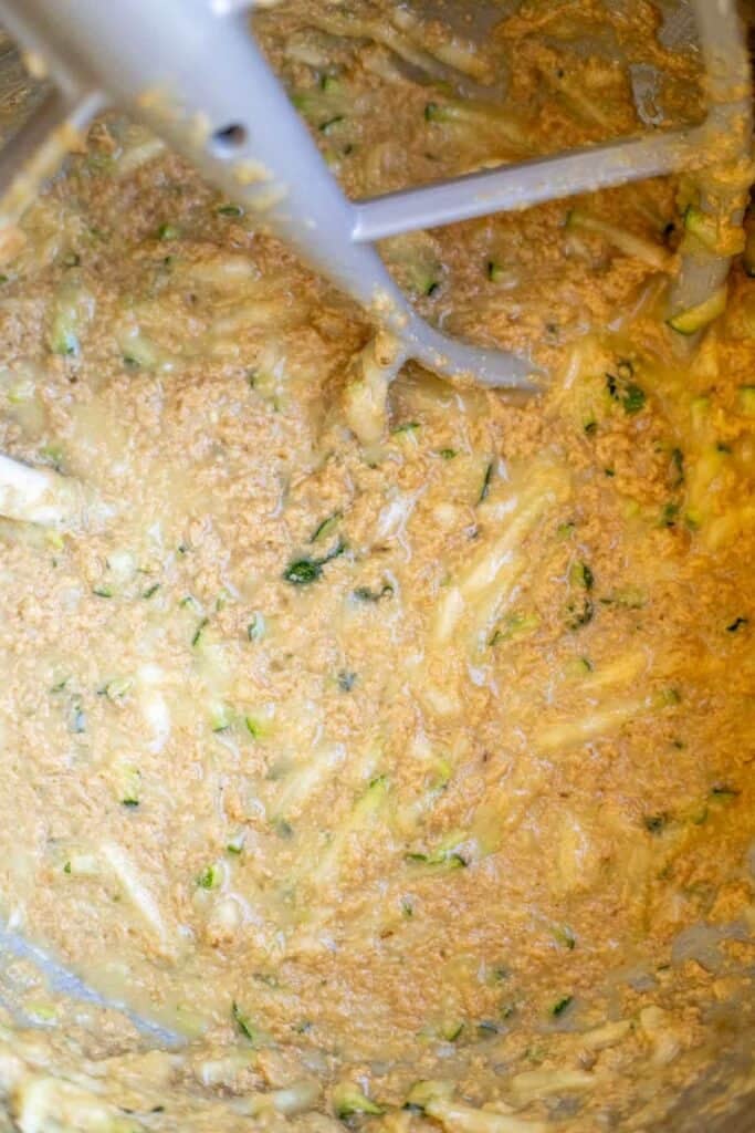 Zucchini and other wet ingredients mixed together in the bowl of a stand mixer with a paddle attachment.