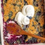 A casserole dish of blueberry cobbler with a wooden spoon laying on top with two dollops of whipped cream.