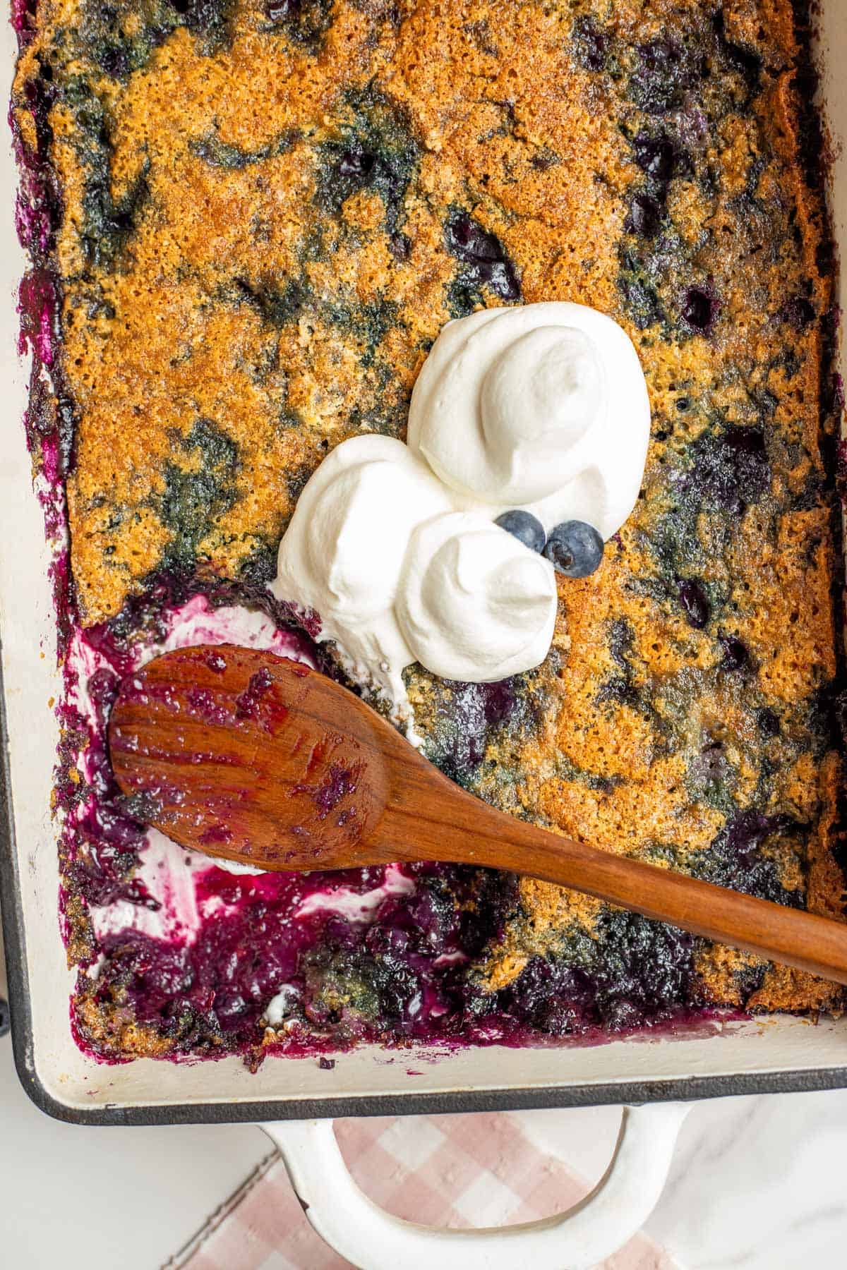 A casserole dish of blueberry cobbler with a wooden spoon laying on top with two dollops of whipped cream.