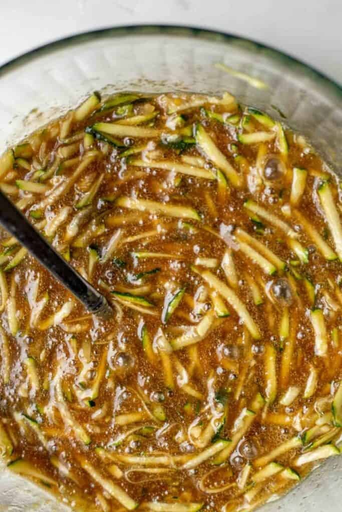 Shredded zucchini mixed into wet ingredients in a glass bowl using a silver spoon.