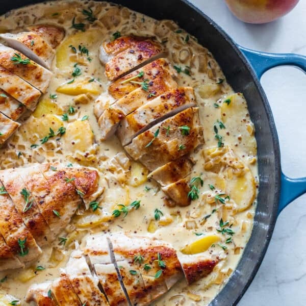 A large cast iron skillet on a countertop filled with chicken breasts sitting in a cream sauce with sliced peaches.