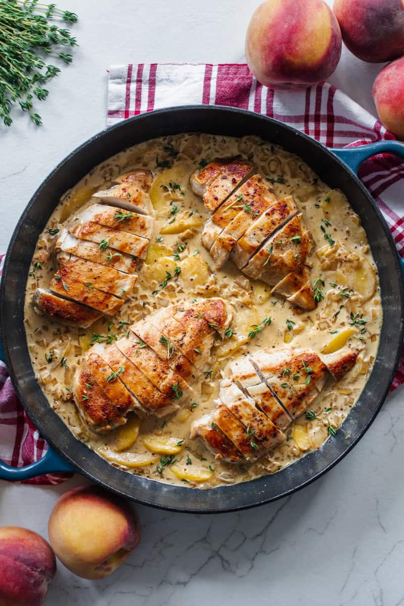 A skillet with four chicken breasts in a cream sauce on a counter top with peaches surrounding it.
