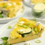 A slice of zucchini frittata topped with herbs on a white plate with more frittata slices in the background.