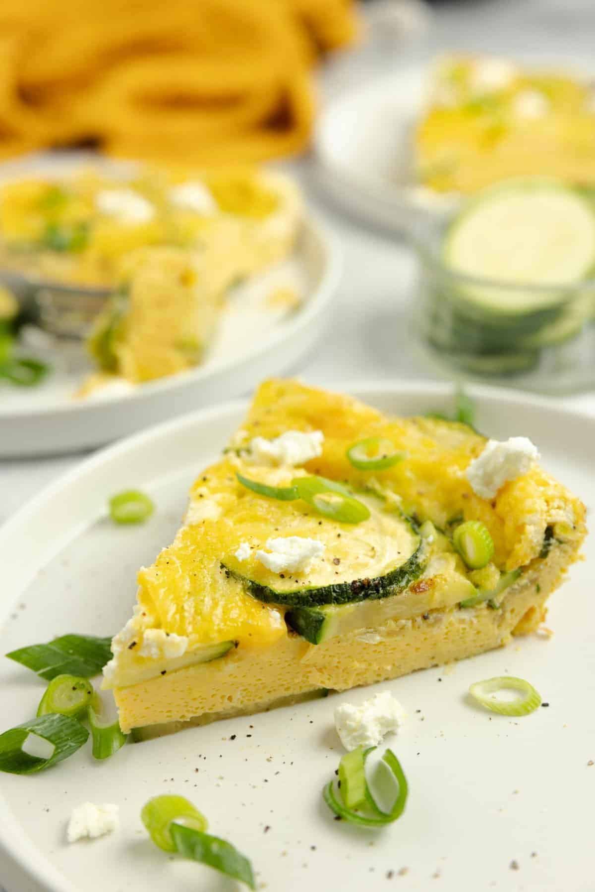 A slice of zucchini frittata topped with herbs on a white plate with more frittata slices in the background.