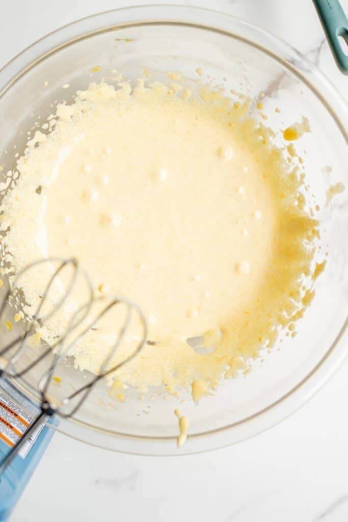 A handheld mixer with a whisk attachment above a glass bowl with eggs and sugar.