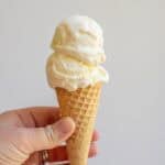 A hand holding an ice cream cone with two scoops of vanilla ice cream.
