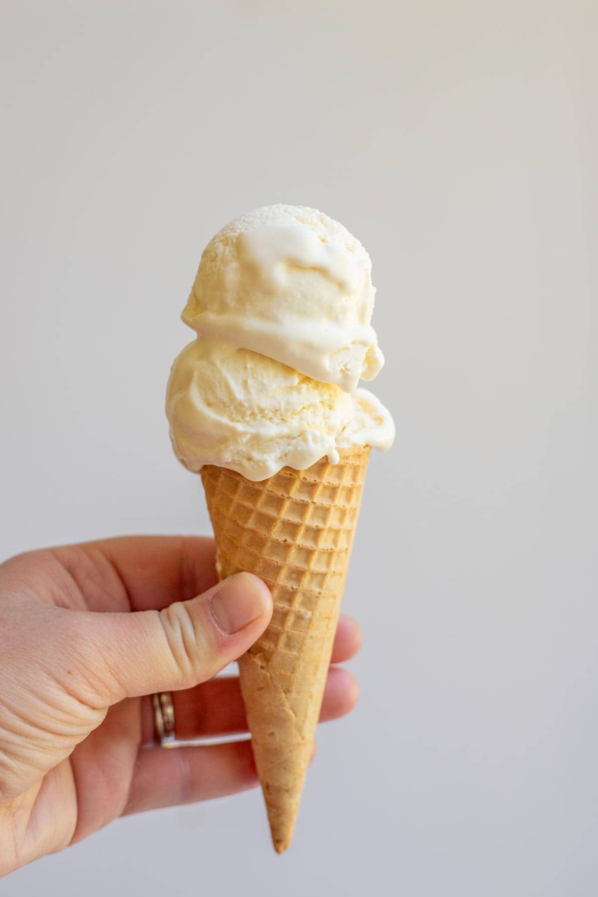 A hand holding an ice cream cone with two scoops of vanilla ice cream.