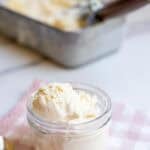 A scoop of ice cream in a glass jar with a loaf pan full of ice cream in the background.
