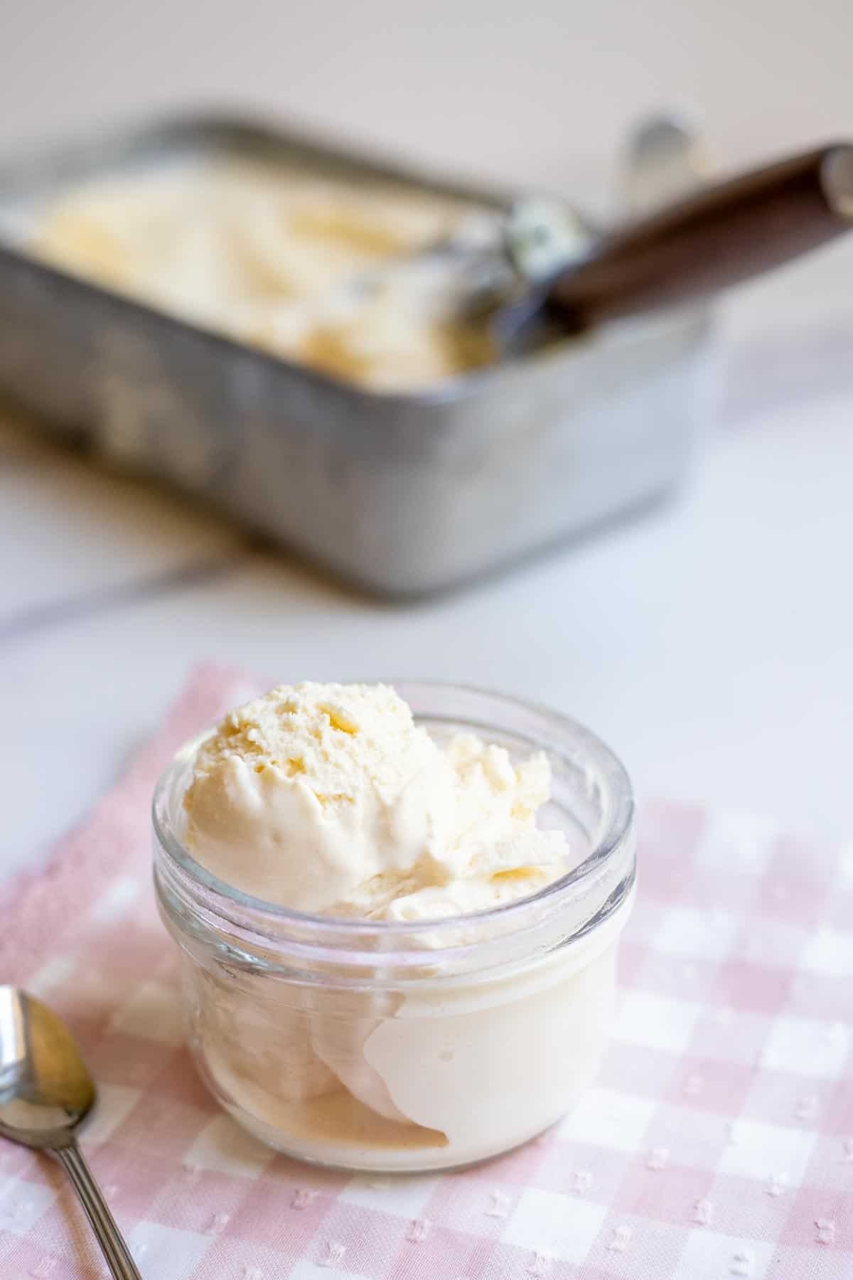A scoop of ice cream in a glass jar with a loaf pan full of ice cream in the background.