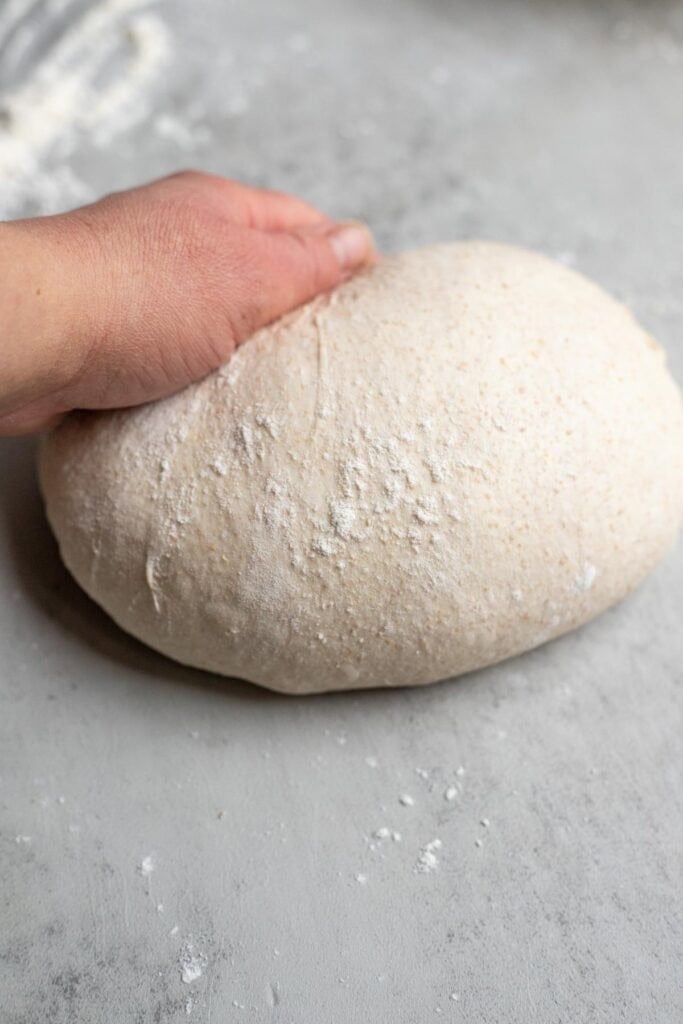 A hand shaping a ball of dough on a clean work surface dusted with flour.