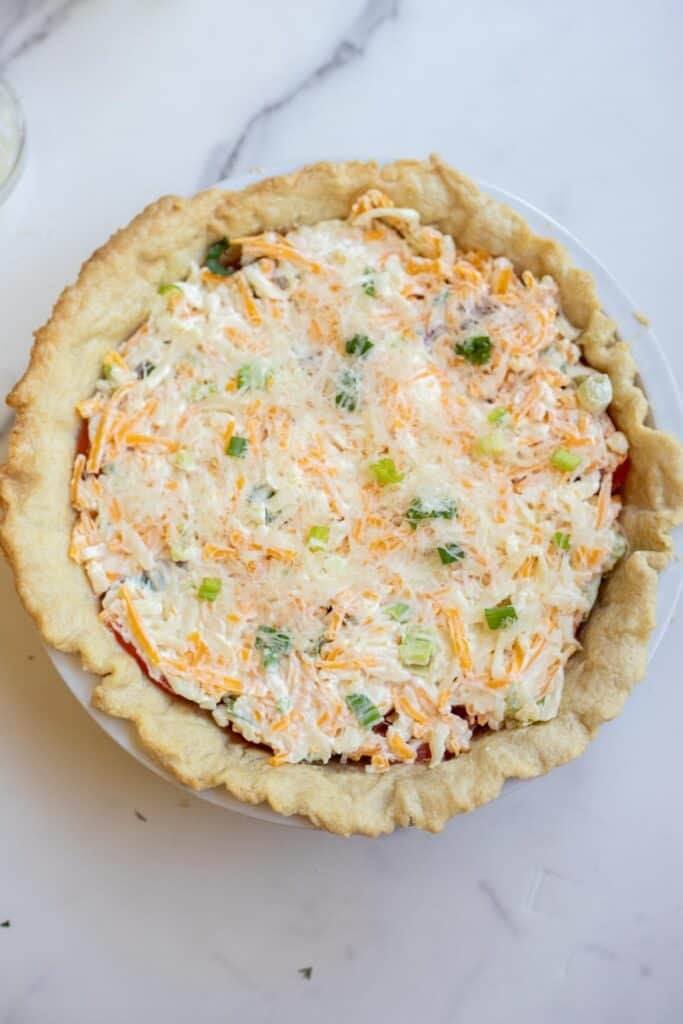 A cheese and mayo mixture on top of slices of tomato in a prebaked pie crust sitting on a countertop.
