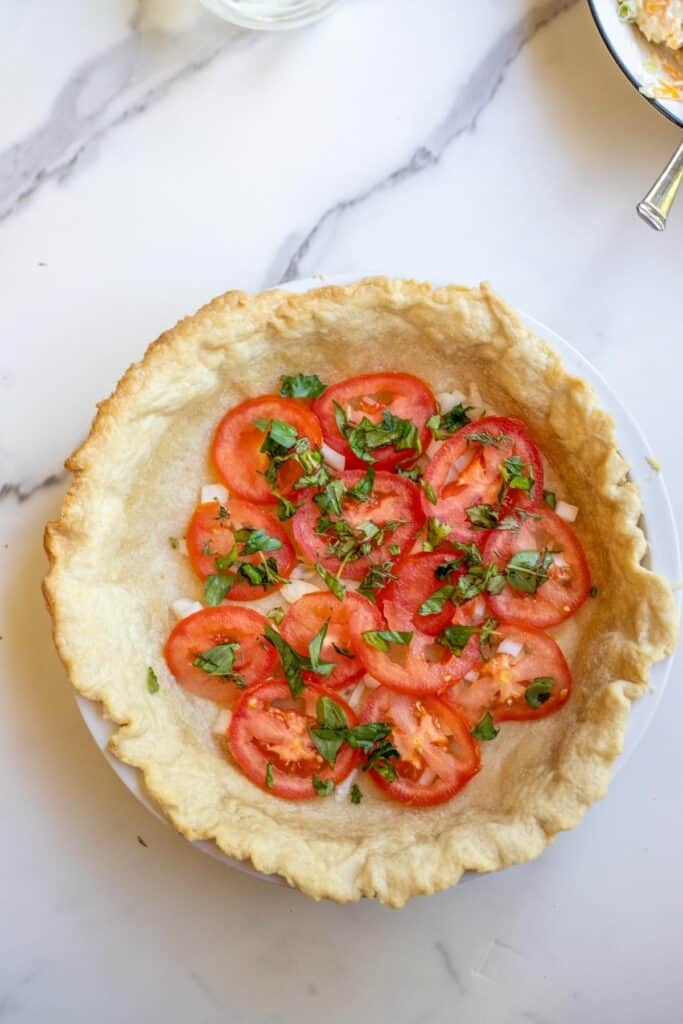 Slices of tomatoes layered into a pie crust in a pie dish with herbs and onion sprinkled on top.