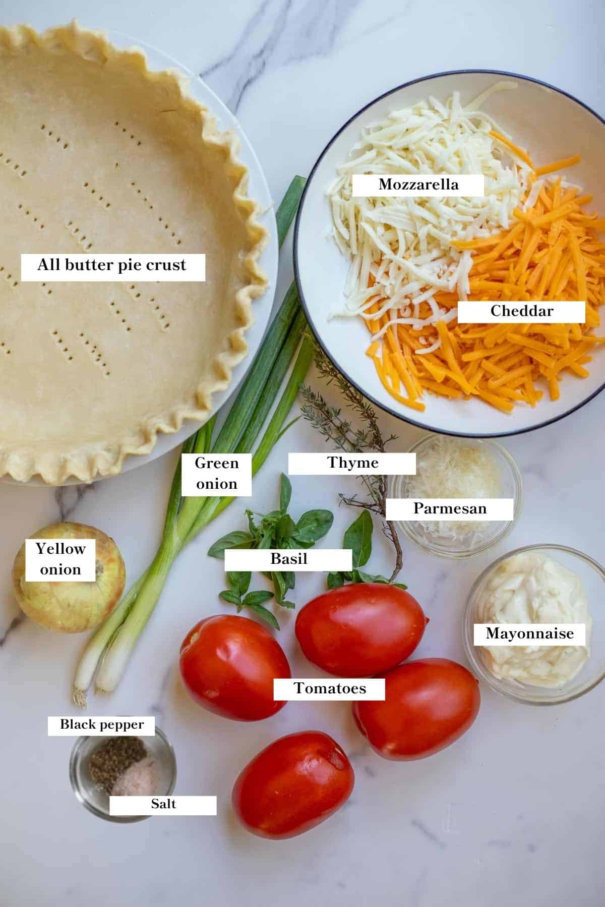 Bowls of ingredients and a pie crust in a pie pan all on a countertop, along with onions, tomatoes, and herbs.