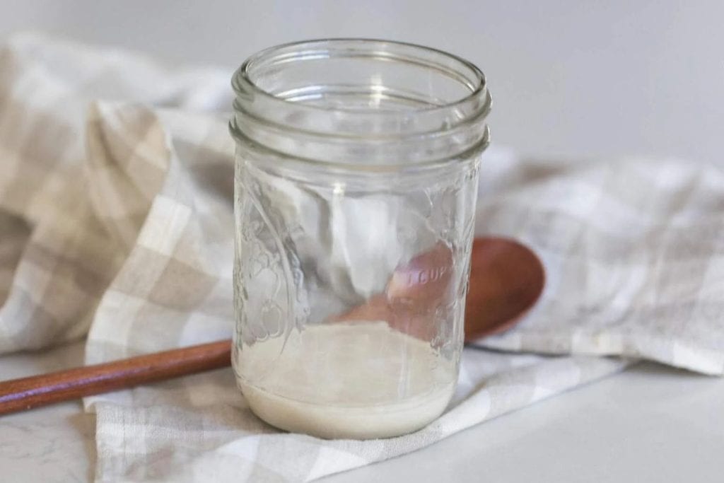 A small jar with thawed sourdough starter in the bottom next to a wooden spoon and tea towel.