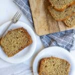 two slices of banana bread on white plates with a cutting board and loaf of banana bread beside it.