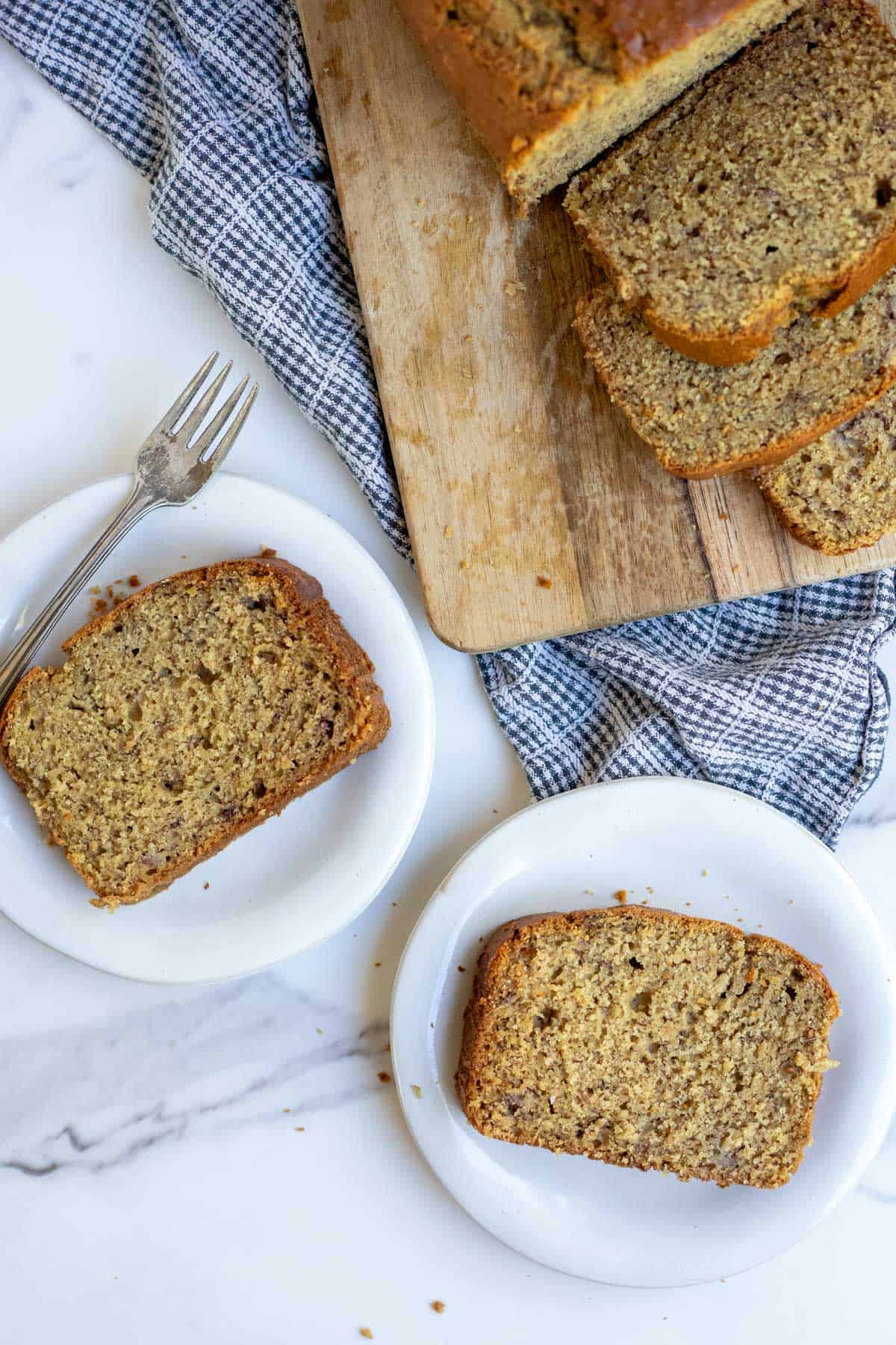 two slices of banana bread on white plates with a cutting board and loaf of banana bread beside it.
